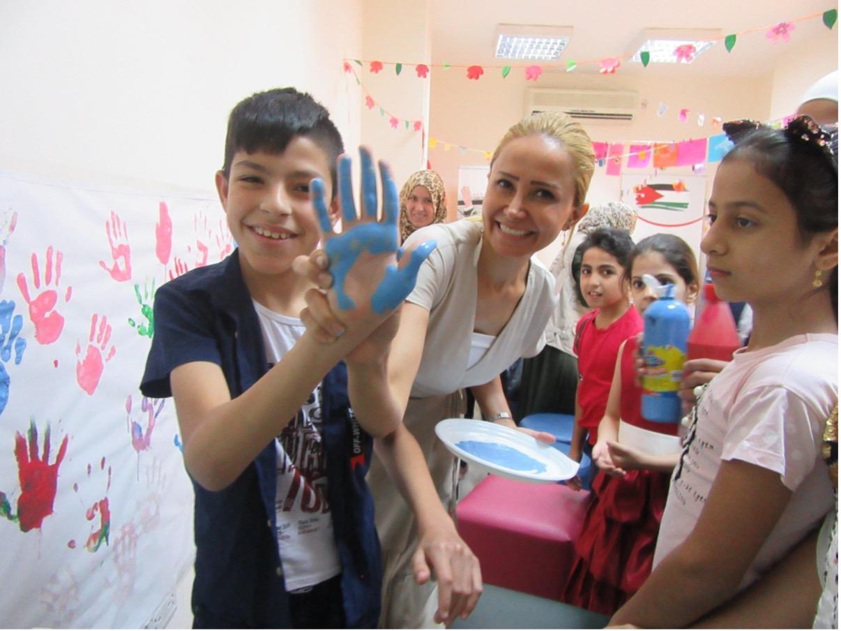 Group of children smiling with Wings of Peace’s Director and Psychologist, Dr. Niveen Abuzaid. This photo shows Dr. Niveen holding a young boy’s hand up covered in blue paint with other children smiling in the background in the expressive arts center. 