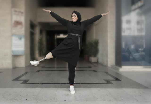 Eman Al-Shayyab smiling while standing on one leg with her arms raised in front of Polus' and ADT's rehabilitation center in Amman, Jordan. Her raised leg is a prosthetic provided to her by Polus and ADT.